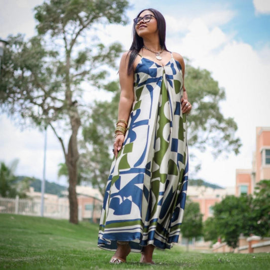 A woman standing in a grassy area wearing a geometric patterned maxi dress with a V-neck and slender shoulder straps, with trees and buildings in the background.