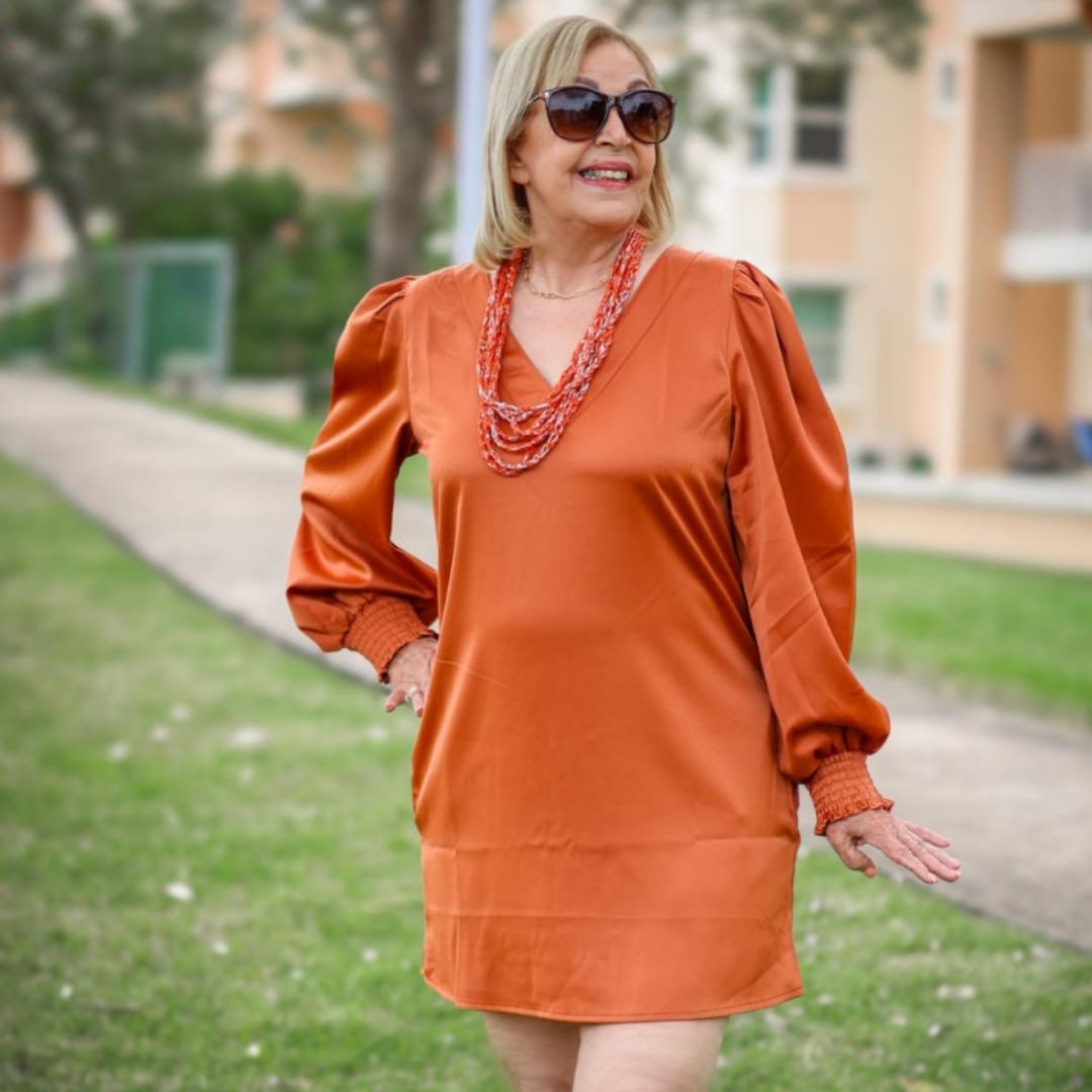 A woman standing on a pathway wearing an orange V-neck long sleeve dress with sunglasses and a necklace.