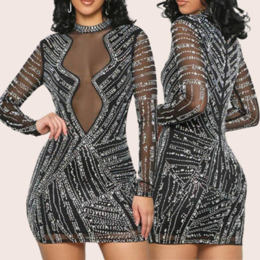A model wearing a sleeveless, sheer, black mini dress with rhinestone embellishments and a zipper at the back.