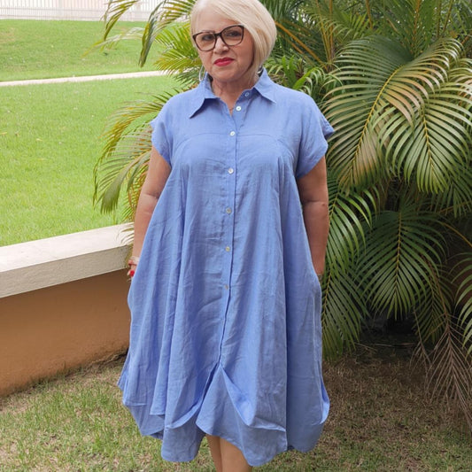 A woman standing by a tropical plant, wearing a blue linen dress with short sleeves, a round neckline, and button details. The dress is fitted at the top and waist and flares out in the skirt.