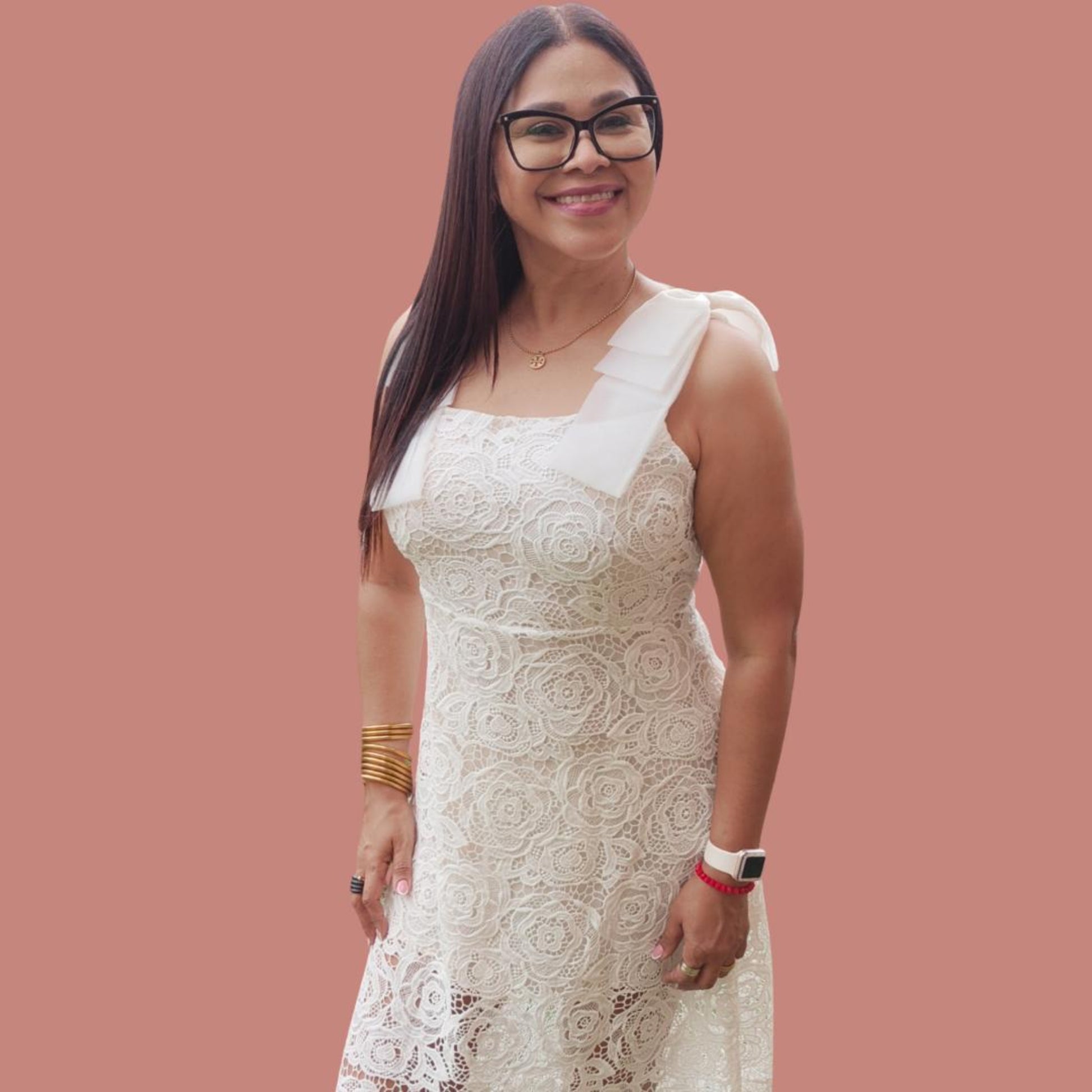 A woman wearing a sleeveless, white, embroidered dress with asymmetrical hem and lace details, standing against a pink background.