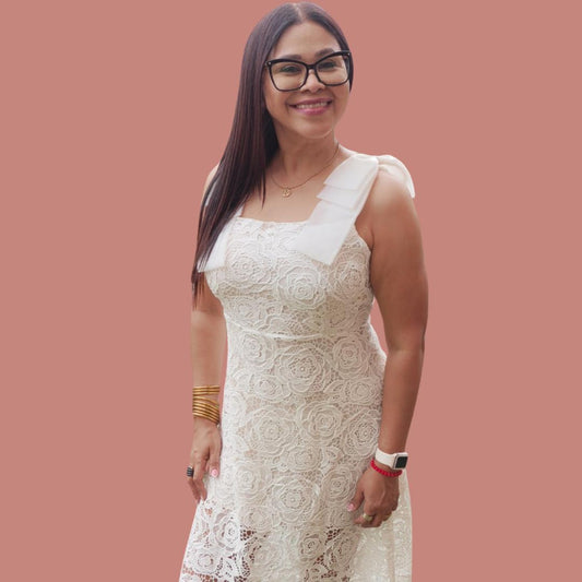 A woman wearing a sleeveless, white, embroidered dress with asymmetrical hem and lace details, standing against a pink background.