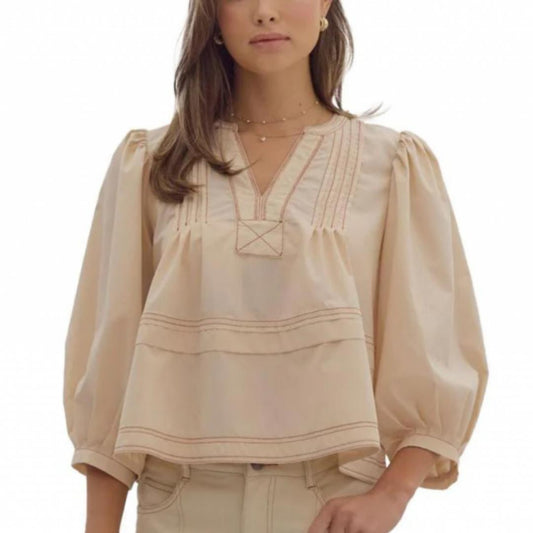 A woman wearing a beige, V-neck, short-sleeved pleated blouse with contrast stitch detail.