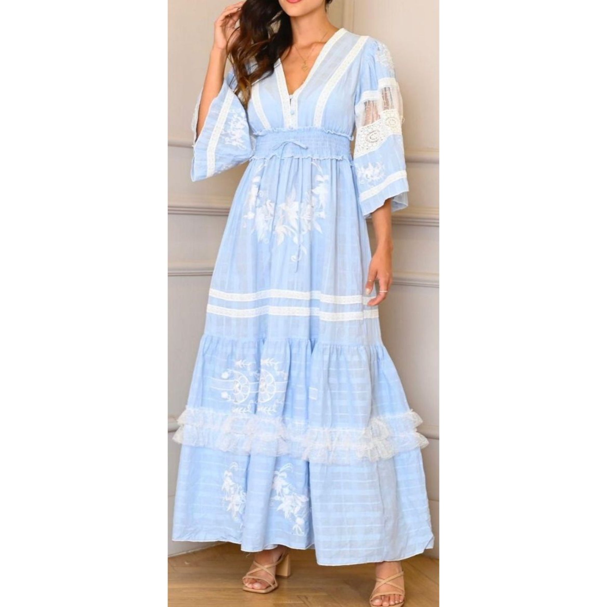 A woman wearing a flowing, floor-length maxi dress in a soft pastel blue color with white floral embroidery, a V-neckline, three-quarter length sleeves with sheer lace inserts, and a cinched waist.
